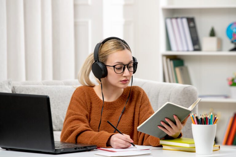Studentka w okularach i swetrze uczy się przy komputerze, obok niej leży otwarta książka.